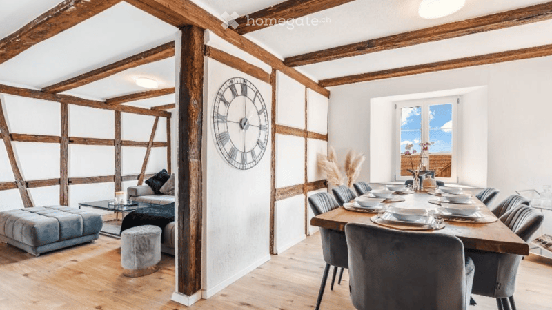 Open-plan living and dining area with exposed wooden beams, a large wall clock, a grey sofa, ottoman, and a long wooden dining table set for eight.