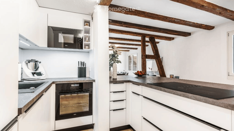 Modern kitchen with white cabinets, stainless steel appliances, exposed wooden beams, and a central island with a black induction stove. Includes a blender, knife block, vase with flowers, and a bowl of fruit on the counter.