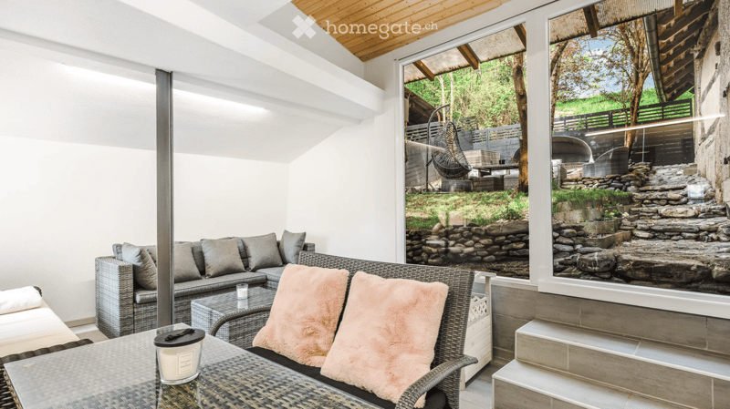 A modern, well-lit living space with a grey wicker sofa, pink throw pillows, a glass coffee table, and a candle on a wicker side table. Large windows offer a scenic view of a landscaped garden with stone steps and outdoor seating.