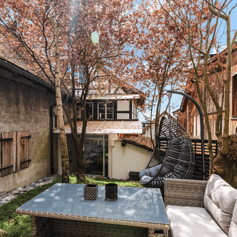 A serene garden area with a wicker hanging chair, a glass-top table with a small decorative box and a mug, and a wicker sofa with cushions. The space is surrounded by trees with autumn foliage and a traditional house in the background.