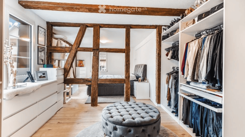 Cozy bedroom with exposed wooden beams, a large bed, a grey tufted stool, a built-in wardrobe, and a dresser with a mirror.
