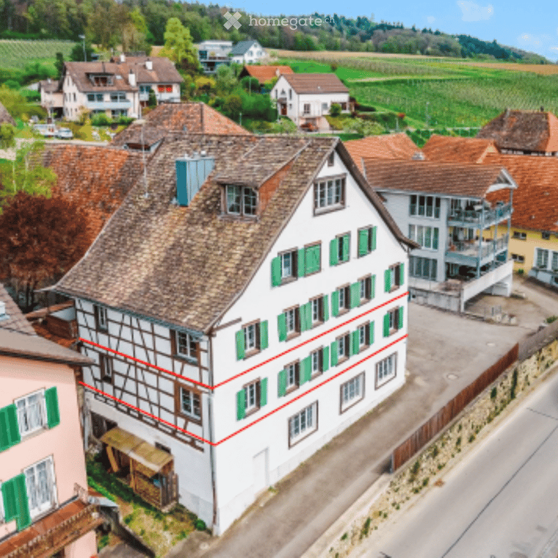 Aerial view of a traditional Swiss house with a pitched roof, green shutters, and a stone facade, surrounded by neighboring houses and green fields.