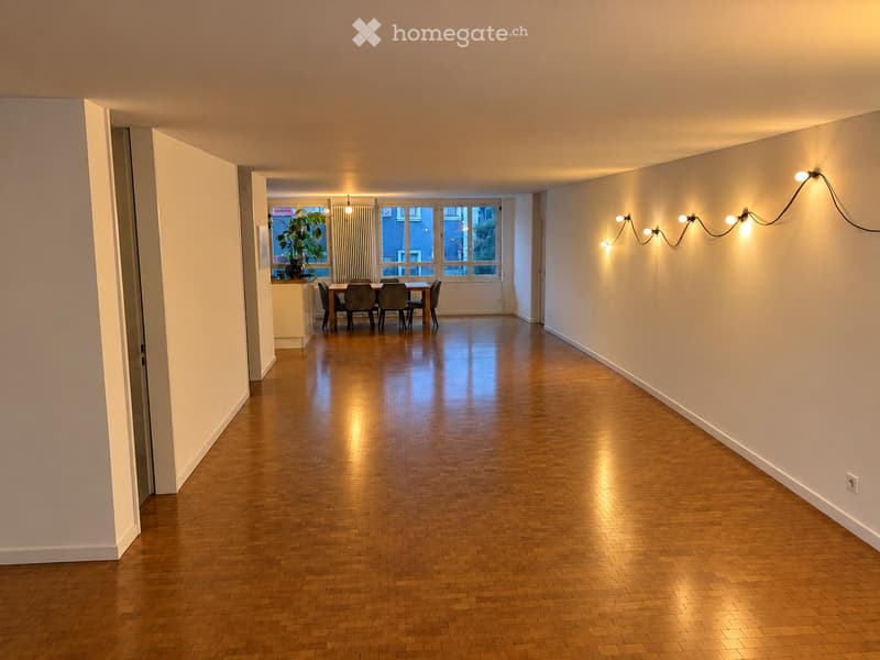 A modern hallway with polished wooden flooring, leading to a dining area with a table and chairs, and a potted plant near a large window.