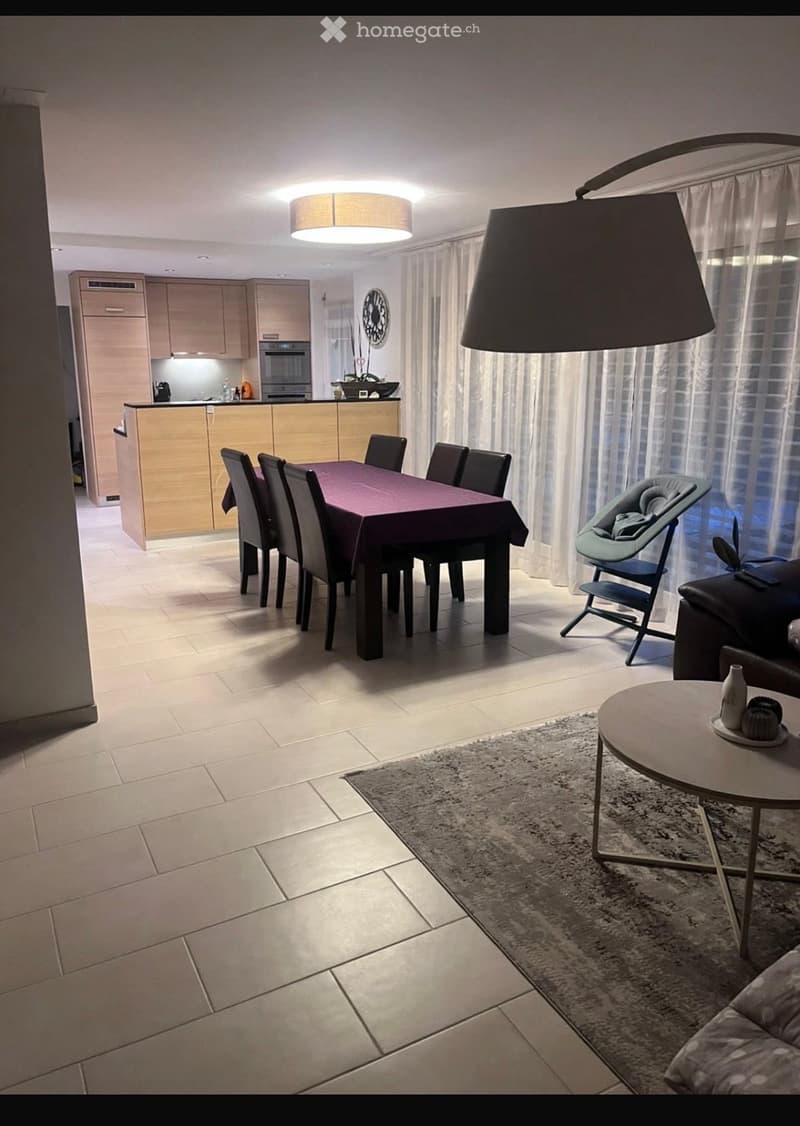 Modern open-plan living and dining area with a sofa, coffee table, dining table with chairs, baby chair, and a kitchen area with cabinets and appliances.