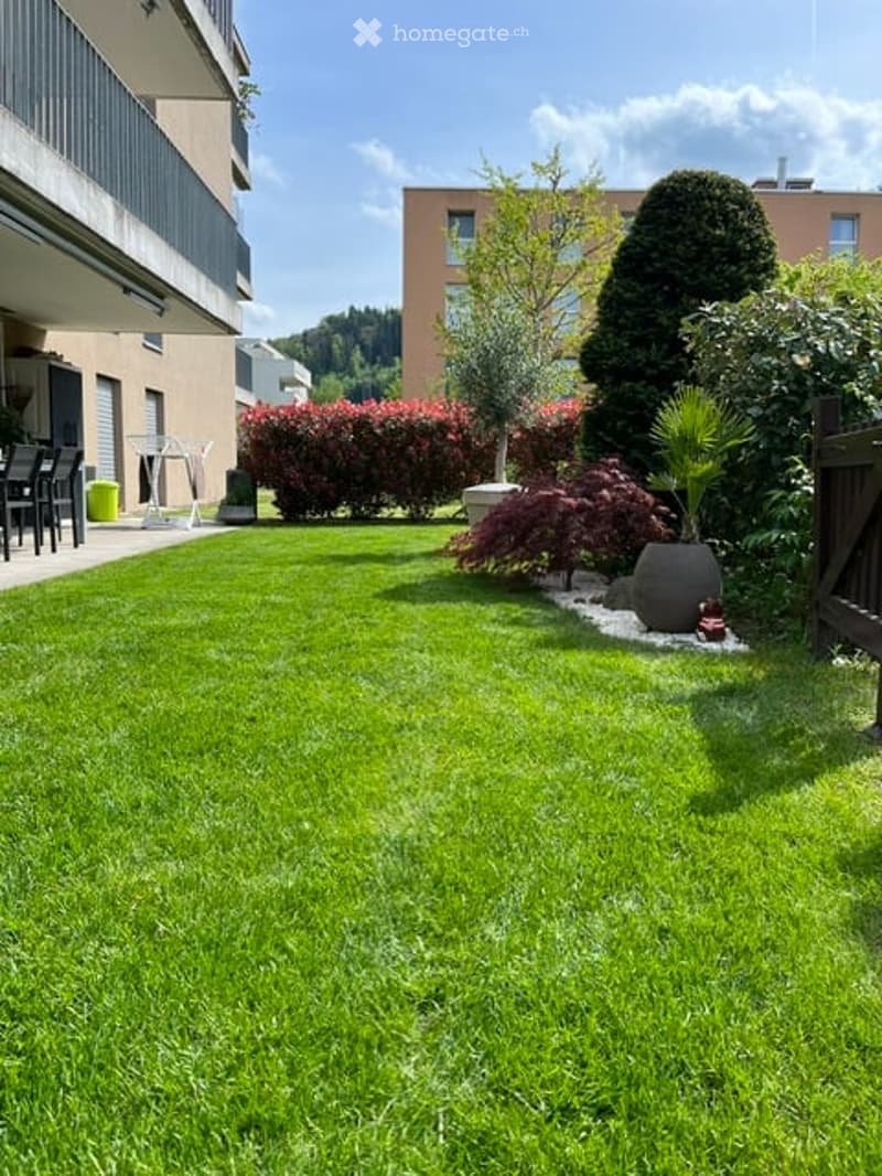 Well-maintained lawn with trimmed hedges, potted plants, and a small seating area near the balcony.
