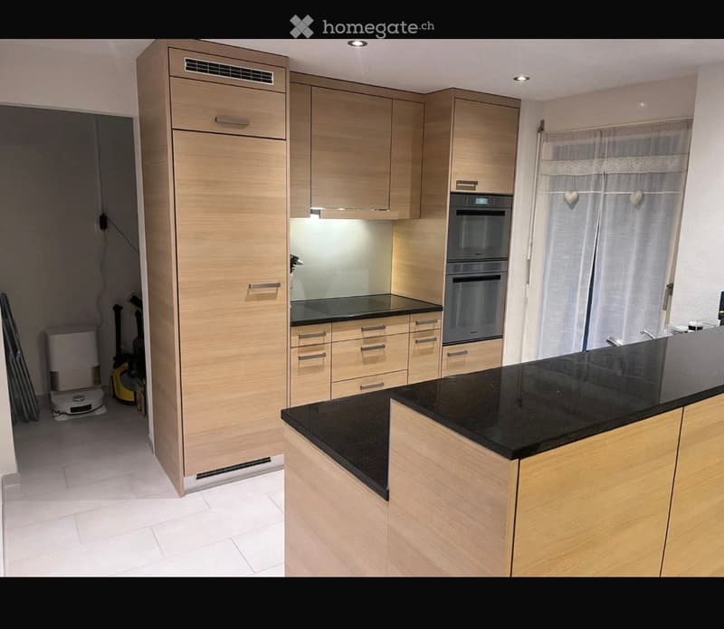 Modern kitchen with light wood cabinets, black countertop, built-in oven, and refrigerator. Features sleek design and ample storage space.