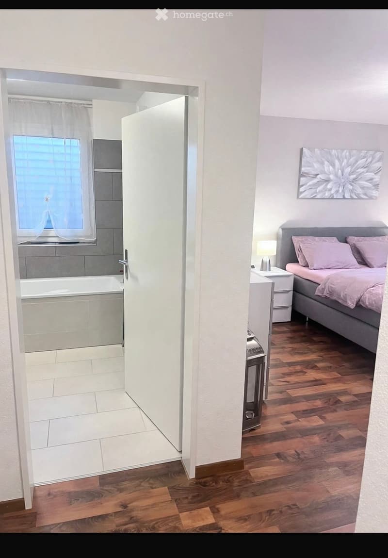 Cozy bedroom with wooden flooring, grey bed with pink bedding, nightstand with lamp, and a picture frame on the wall. An open door leads to a bathroom with a bathtub and window.