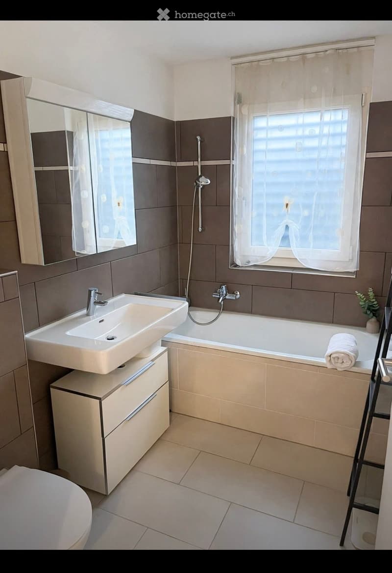 Modern bathroom with a white sink, bathtub, mirror, and a small window with a curtain. The room features beige tiles and a clean, contemporary design.