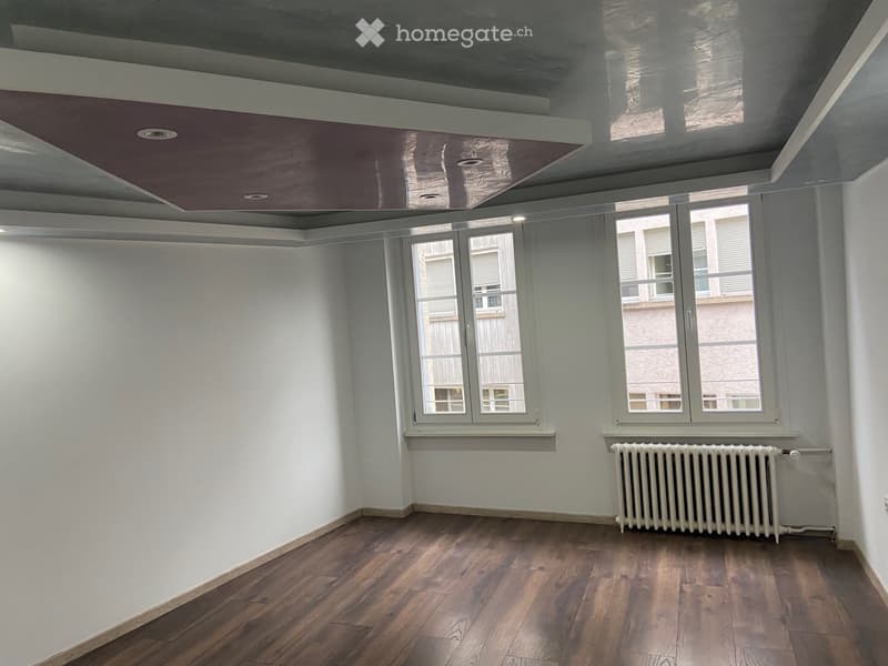 Empty room with wooden wall paneling, white radiator, and large window showing a view of neighboring buildings.