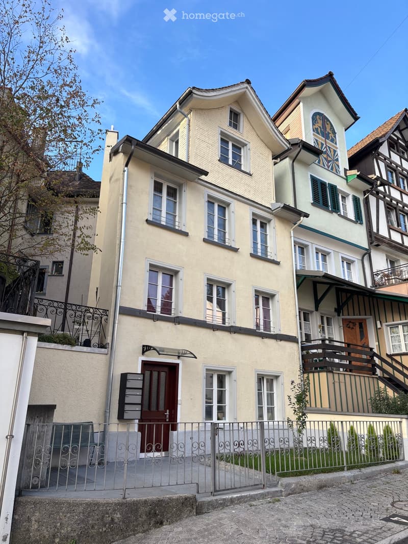 Two-story residential house with beige facade, multiple windows, decorative shutters, and a small balcony. The house features a metal fence and a small garden area in front.