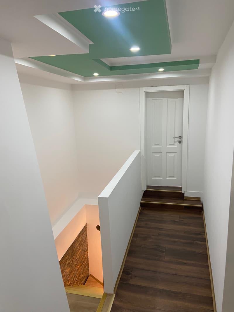 Modern hallway with wooden flooring, white walls, and a green ceiling with recessed lighting. Features a white door at the end and a staircase leading upwards.