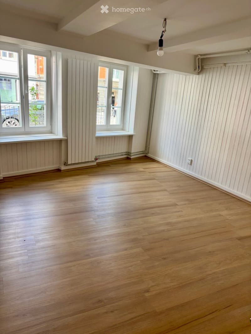 Empty room with wooden flooring, white walls, and large windows with radiators below them.