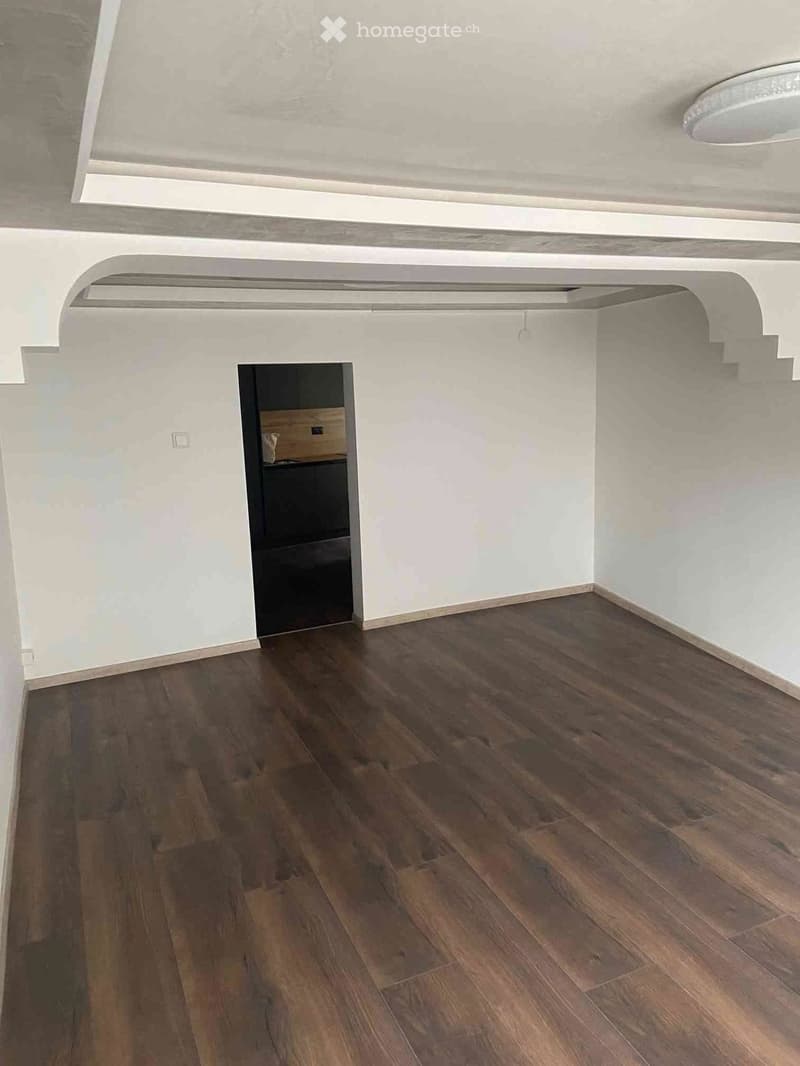 Empty room with wooden flooring and white walls, featuring an arched doorway leading to another space.