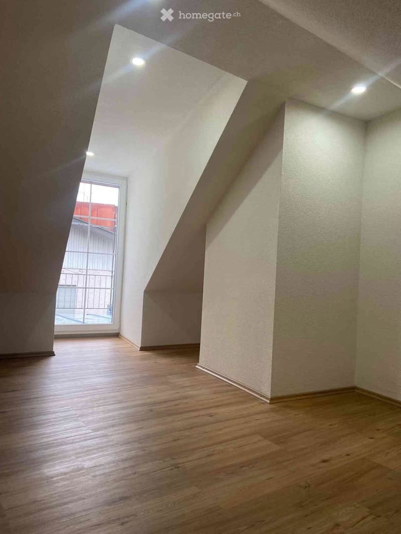 Empty room with wooden flooring, white walls, and a window with bars leading to an outdoor area.