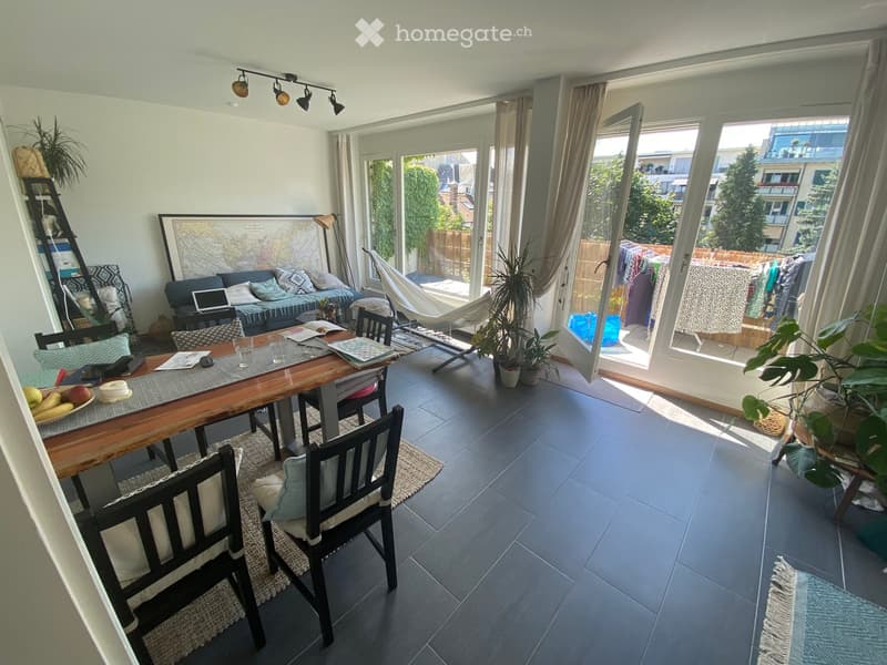 Bright living and dining area with large windows and sliding doors leading to a balcony. Features a sofa, dining table with chairs, laptop, potted plants, and a hammock.