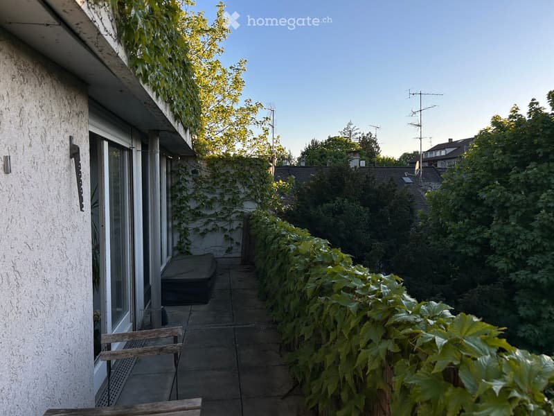 Balcony with wooden bench, covered furniture, and lush greenery, overlooking a garden area with trees and neighboring houses.