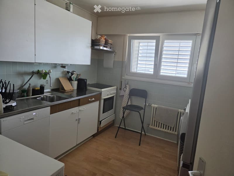A compact kitchen with white cabinets, a dishwasher, oven, sink, and a small window with blinds. Wooden flooring and a radiator with towels are visible.