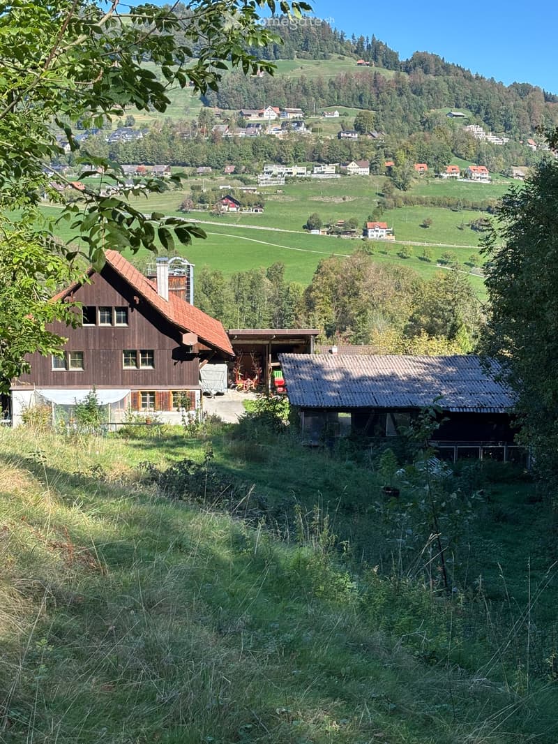 A scenic rural landscape with a wooden house, a small shed, and a view of a valley with scattered houses and green fields.