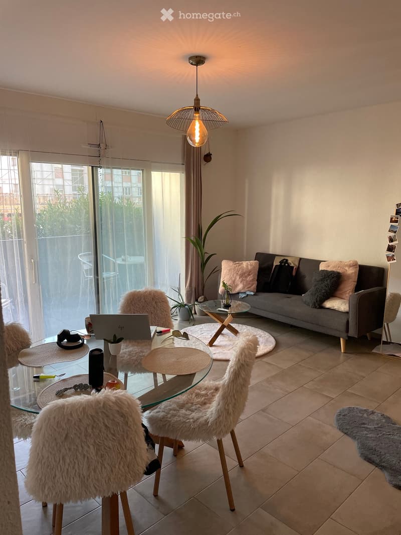 Modern living room with a grey sofa, glass coffee table, fur chairs, and a laptop on a glass dining table. Features a large window with a view of the balcony and outdoor area.