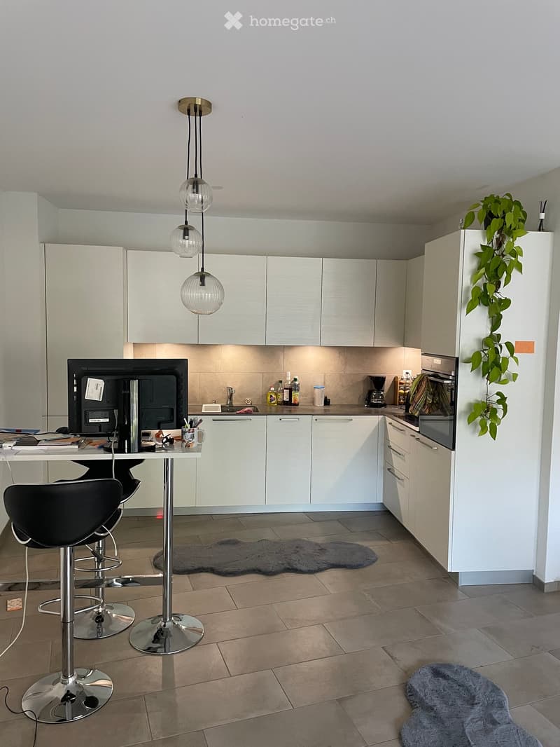 Modern kitchen with white cabinets, stainless steel appliances, a small dining area with stools, and a desk with a computer. Features include a refrigerator with a plant, a coffee maker, and various kitchen items on the counter.