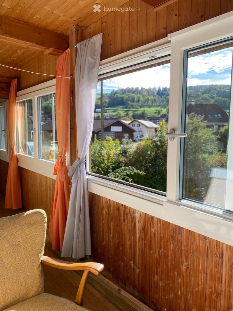 Cozy room with wooden walls and ceiling, large windows offering a scenic view, and a single armchair.