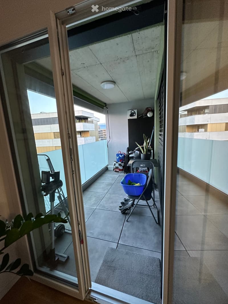 Balcony with tiled floor, glass railings, and a view of the building exterior. Includes a folding chair, a blue laundry basket, a small table, and a potted plant.