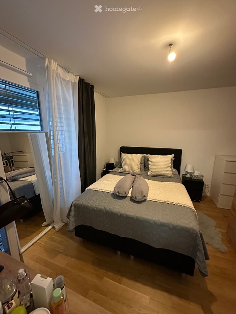 Cozy bedroom with a double bed, nightstands, lamps, and a large window with curtains. Wooden flooring and a small rug near the bed.