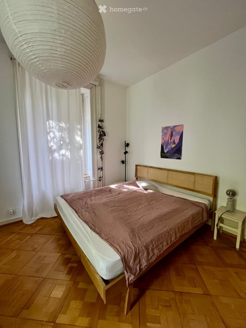 Cozy bedroom with wooden flooring, a large bed with a brown duvet, a nightstand with a lamp, and a large window with white curtains.
