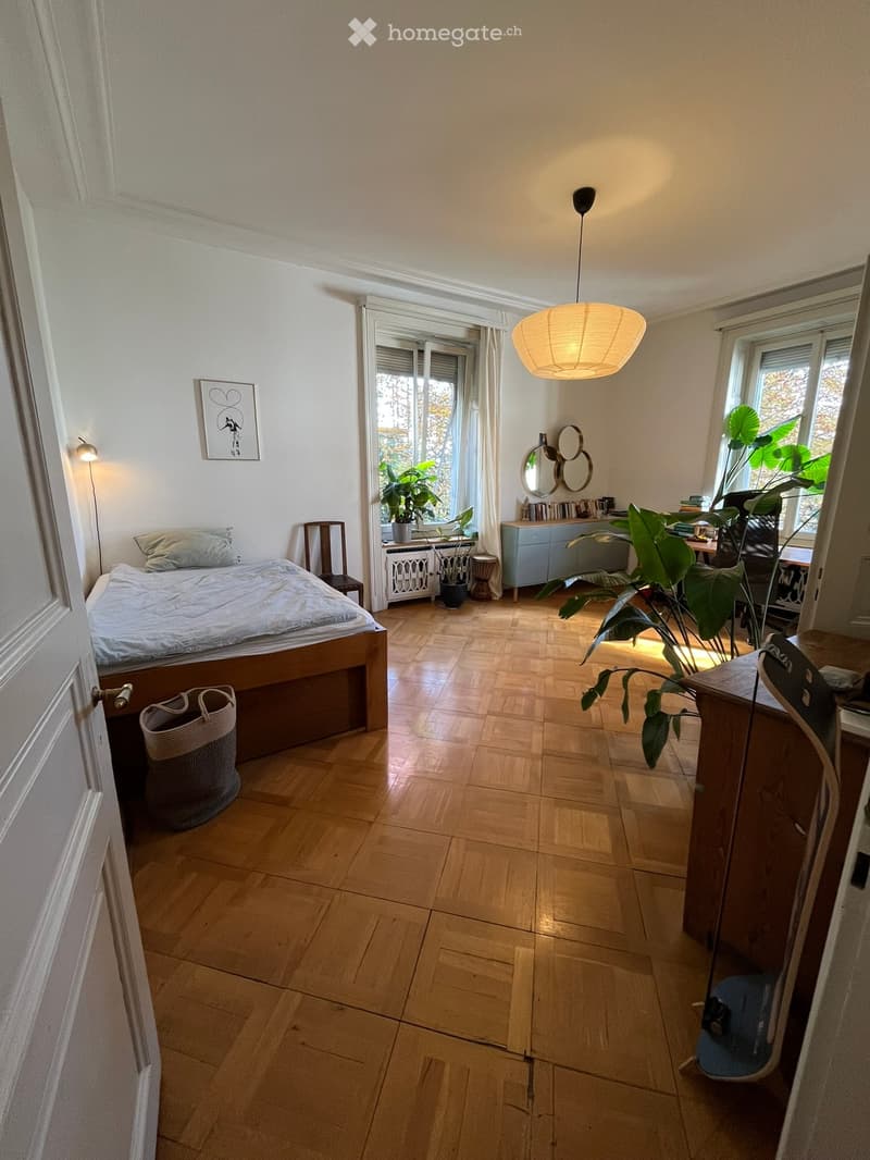 Bright bedroom with wooden flooring, large windows, bed with pillows, nightstand, chair, mirror, cabinet, potted plants, picture frame, and a hanging lamp.