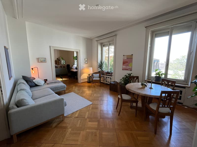 Bright living room with wooden flooring, large windows, a grey sectional sofa, a round wooden dining table with chairs, a side table with a lamp, and a small workspace area with a desk and chair.