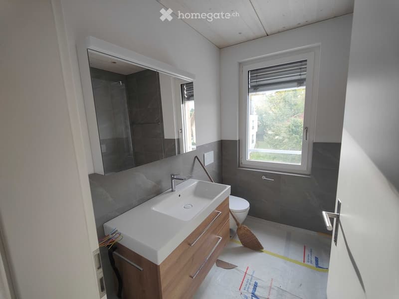 Modern bathroom with a white sink, wooden cabinet, large mirror, and a toilet. The walls are tiled in grey, and there is a window with blinds allowing natural light.