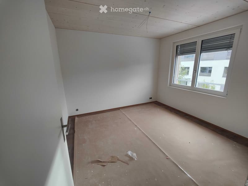Empty room with bare walls, wooden baseboard, and a window with blinds. The floor is unfinished and shows some construction debris.