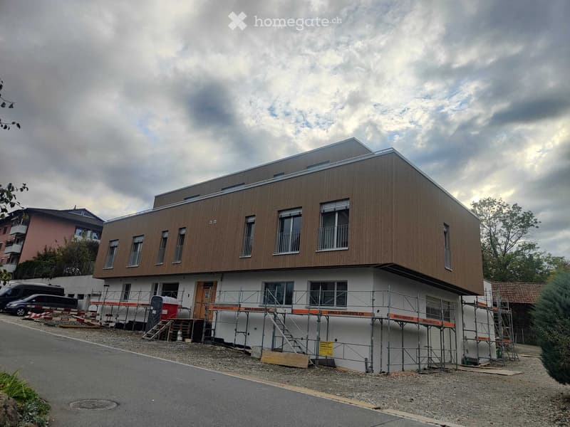 Modern two-story building under construction with wooden facade and white lower section, featuring multiple windows and a flat roof.