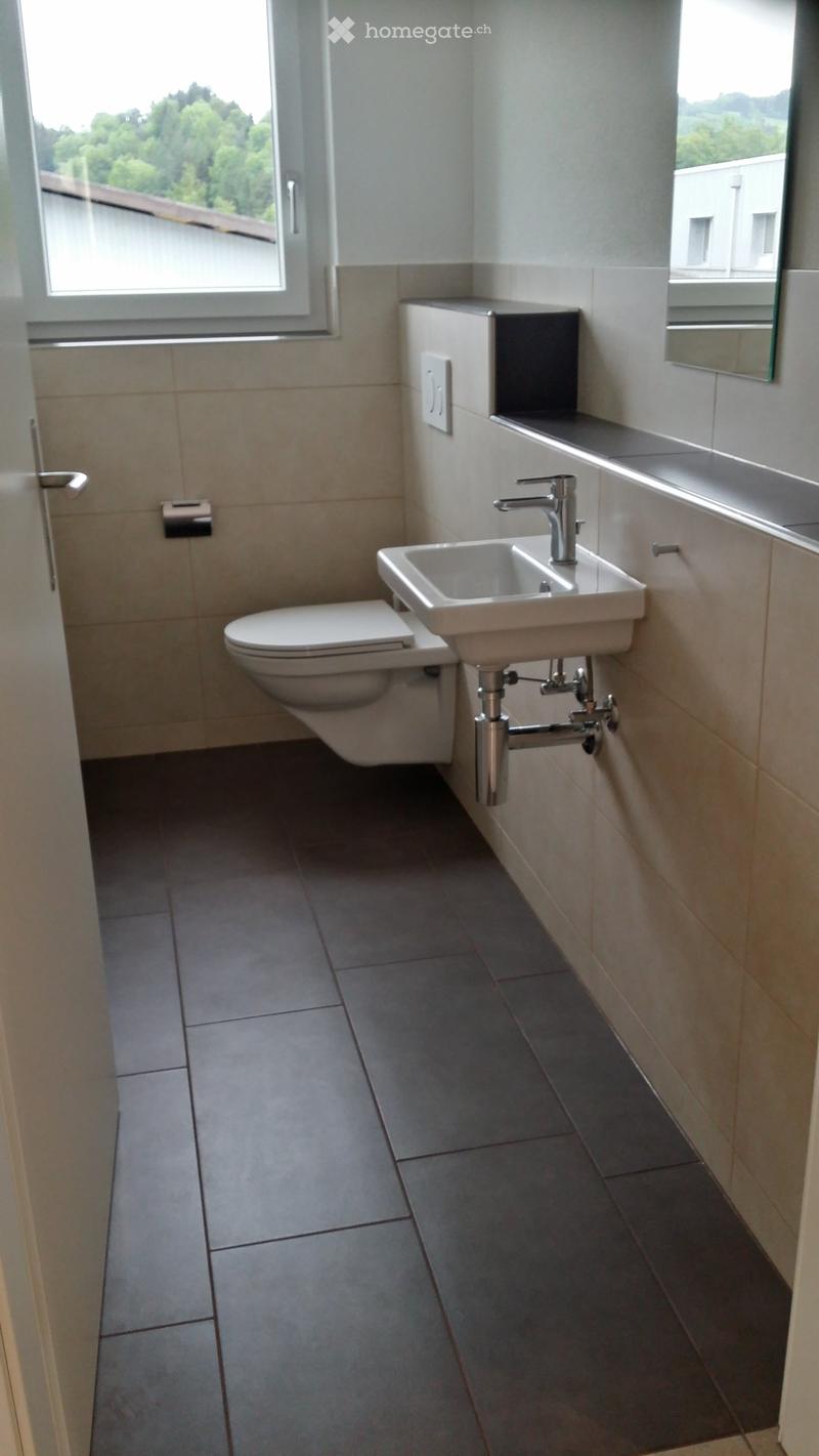 Modern bathroom with a toilet, sink, and mirror. The sink has a modern chrome faucet, and the walls are tiled with light beige tiles. The floor is covered with dark grey tiles.