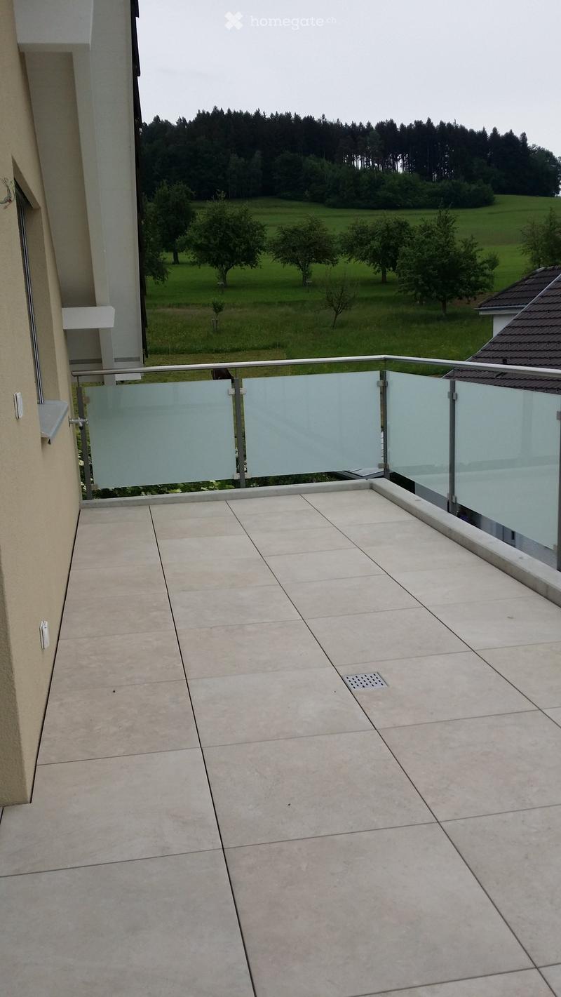 Balcony with tiled floor, glass railing, and scenic view of a green field and trees.