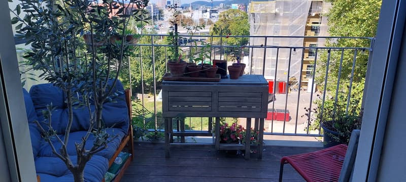 Balcony with wooden decking, metal railing, small sofa, table, and potted plants, overlooking a cityscape with trees and buildings.