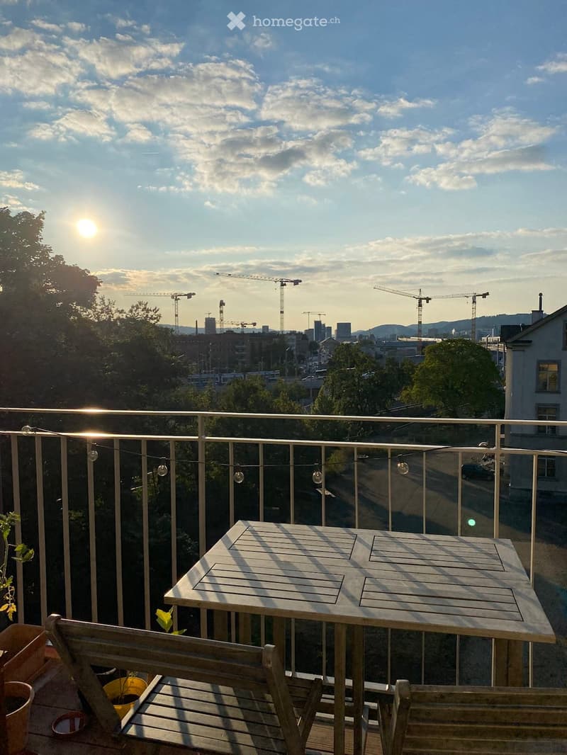 Balcony with a wooden table, bench, and chairs, overlooking a cityscape with cranes and buildings.