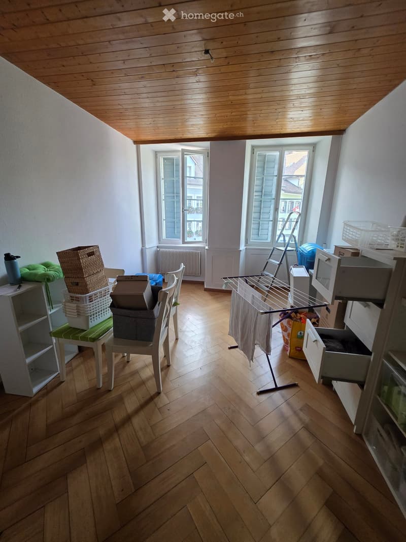 A room with wooden flooring and white walls, featuring a chair, shelf, microwave, laundry rack, and a small stool with boxes on it.