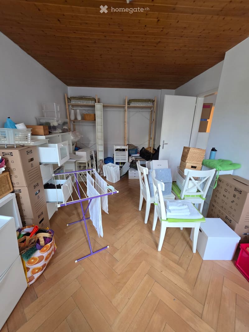 A cluttered room with wooden herringbone flooring, featuring a white chair with folded towels, a purple laundry rack, a refrigerator, microwave, shelves with storage boxes, and various items scattered around.