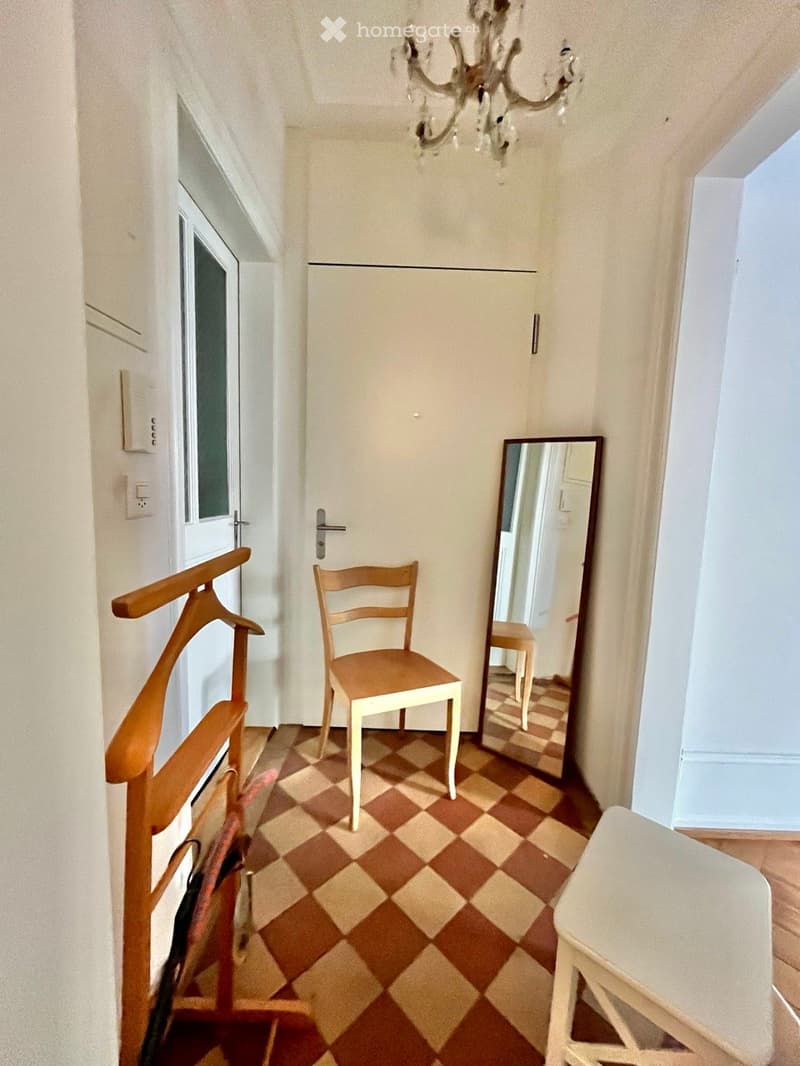 A narrow hallway with checkered flooring, a wooden chair, a large mirror, and a coat rack. The space features white walls and a chandelier hanging from the ceiling.
