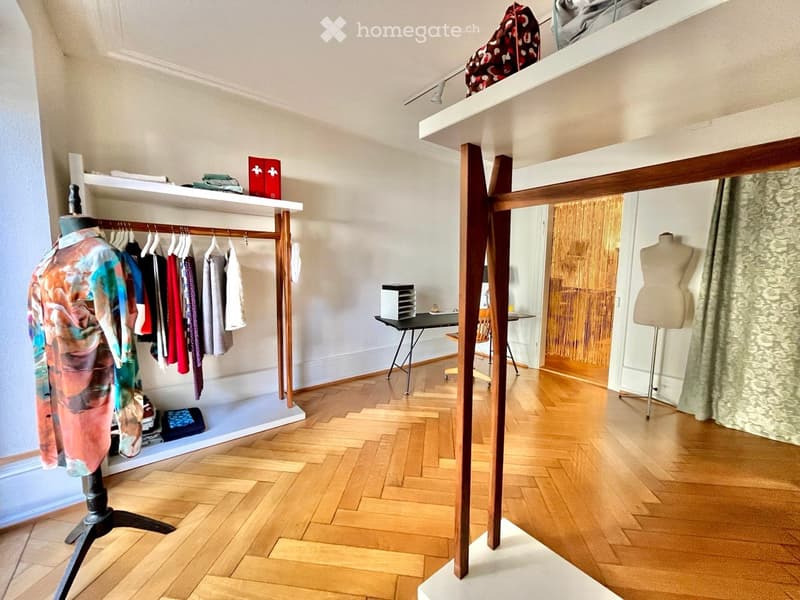A well-lit room with wooden flooring, featuring a clothing rack with various garments, shelves with folded clothes and accessories, a small table with a chair, and a mannequin in the corner.