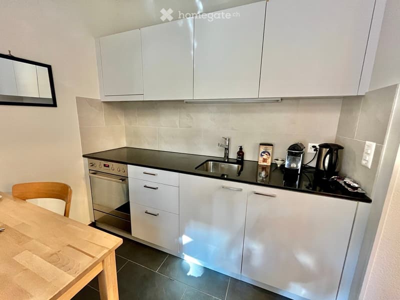 Modern kitchen with white cabinets, black countertop, stainless steel sink, and various appliances including a coffee maker and kettle. A wooden table and chair are visible in the foreground.