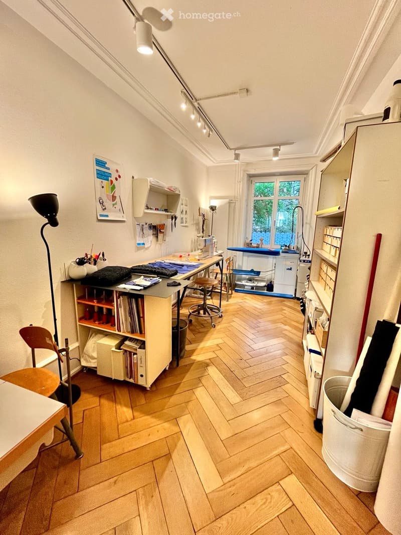 A well-lit room with wooden herringbone flooring, a large window, and various furniture including a chair, table, shelves, and a lamp.