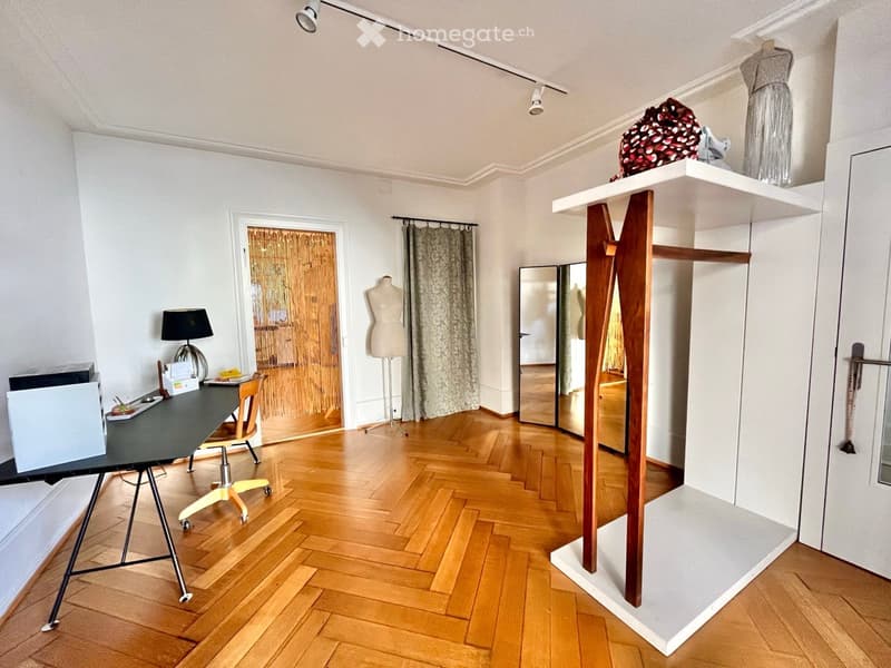 A modern, well-lit room with wooden flooring, a large wooden shelf, a desk with a chair, a lamp, and a mirror. The room features a mannequin and a curtained doorway leading to another space.