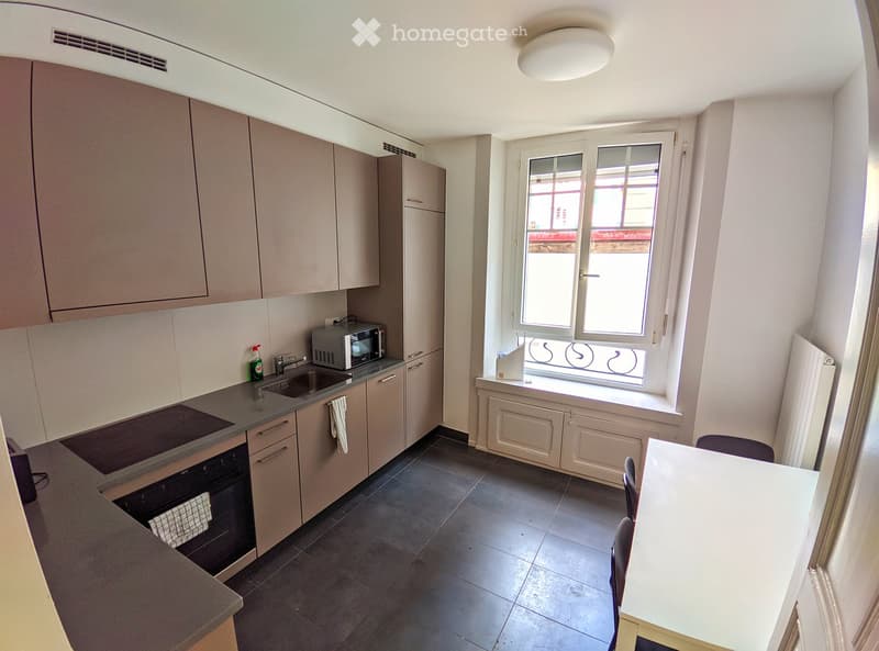Modern kitchen with light-colored cabinets, dark countertops, and a window overlooking the street. Includes a sink, microwave, oven, and a small dining area with a table and chairs.