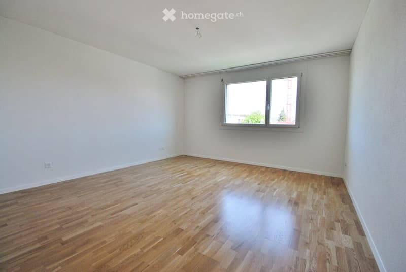 Empty room with wooden flooring and white walls, featuring a large window with a view of trees and a building outside.