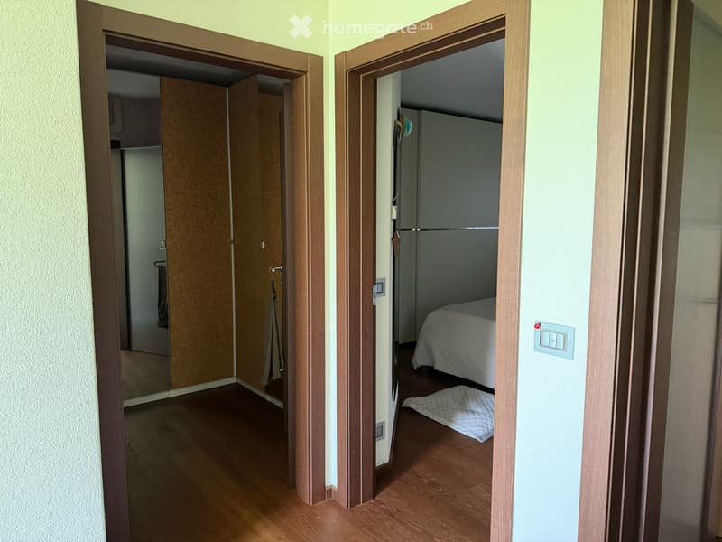 Hallway with wooden flooring, leading to a bedroom and a closet. The bedroom contains a bed with white bedding and a small rug on the floor.
