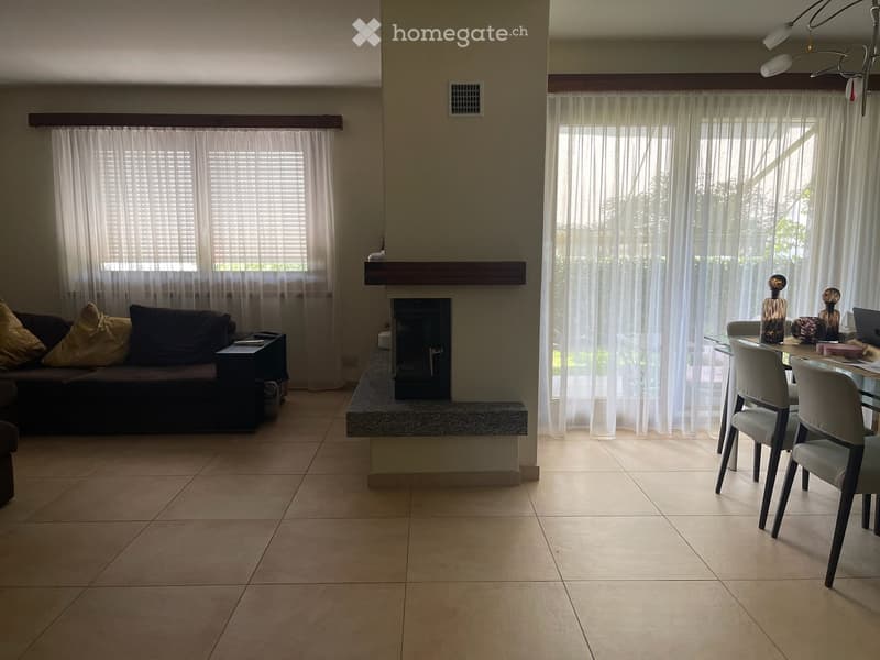 Open-plan living and dining area with tiled flooring, fireplace, large windows with curtains, sofa, dining table, and chairs.