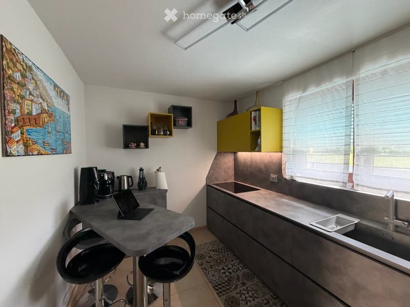 Modern kitchen with grey countertops, yellow cabinets, and a small breakfast bar with stools. Includes a coffee maker, laptop, and a sink with a modern faucet.