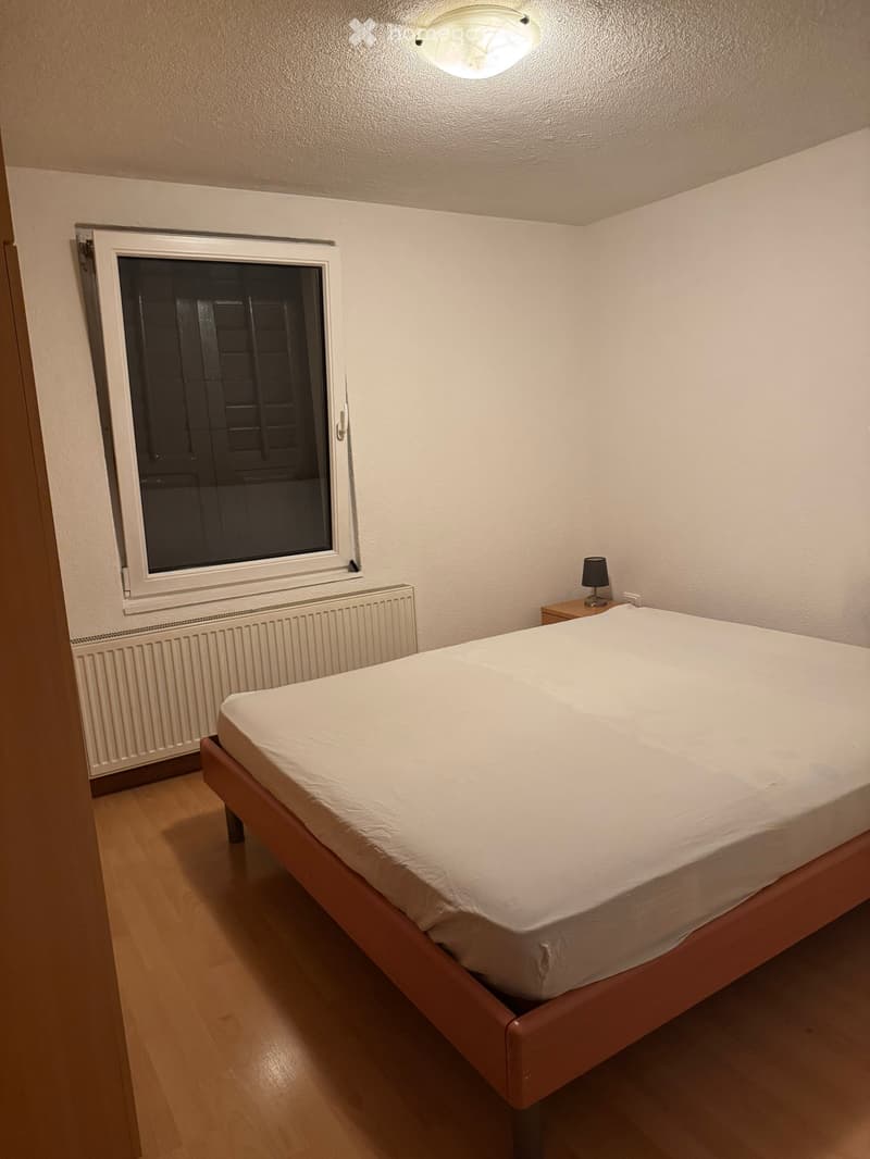 Bedroom with a single bed, nightstand, and lamp. The room features wooden flooring, a radiator, and a window with a dark blind.